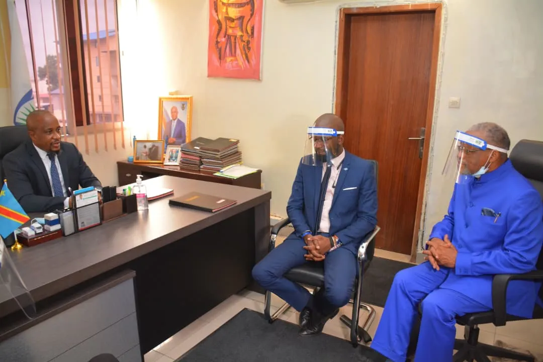 Réunion institutionnelle dans un bureau : trois responsables en entretien, drapeau de la République démocratique du Congo posé sur le bureau.